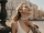 Portrait of beautiful blonde lady in stylish white silk dress, checkered jacket and pearl necklace gently touching neck and posing at city square.