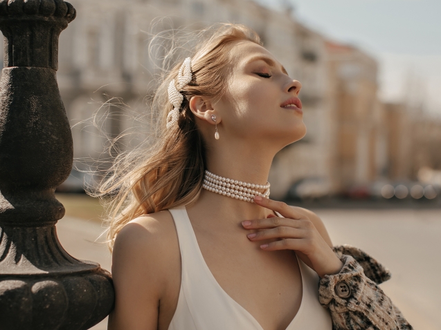 Portrait of beautiful blonde lady in stylish white silk dress, checkered jacket and pearl necklace gently touching neck and posing at city square.