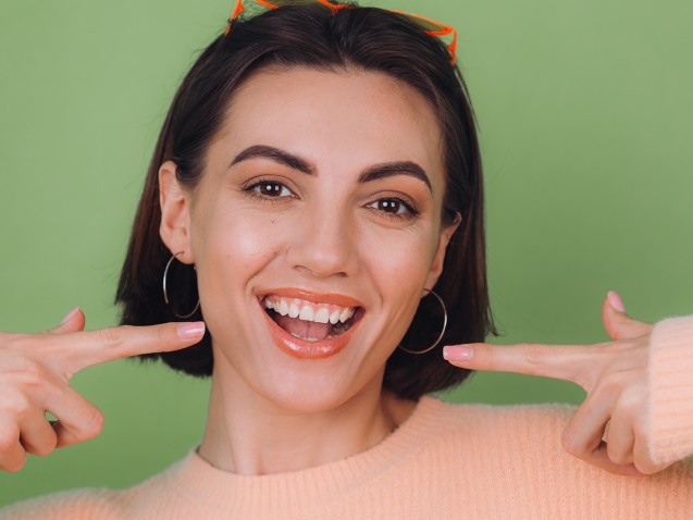 Young stylish woman in casual peach sweater and orange glasses isolated on green olive background positive smiling pointing on white teeth with index fingers copy space