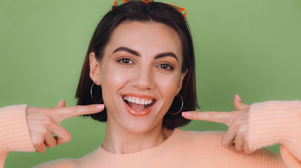 Young stylish woman in casual peach sweater and orange glasses isolated on green olive background positive smiling pointing on white teeth with index fingers copy space